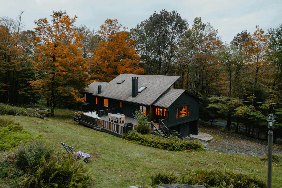 A dark-sided home on a sloping lawn in fall, with lit windows, a furnished deck, and orange and gold trees behind it.