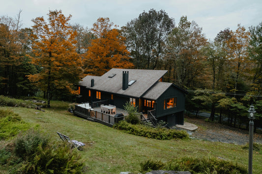 A dark-sided home on a sloping lawn in fall, with lit windows, a furnished deck, and orange and gold trees behind it.