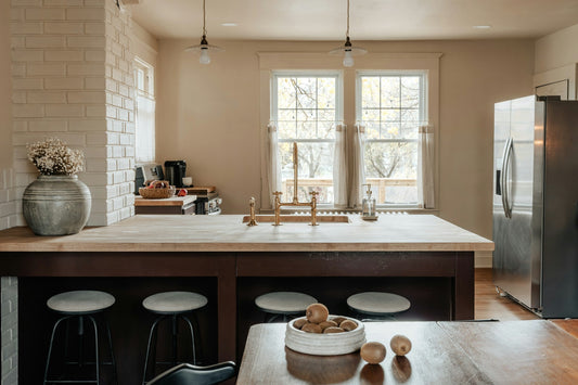 A kitchen in morning light, with a bowl of fruit on the wooden table and coffee set out on the counter.