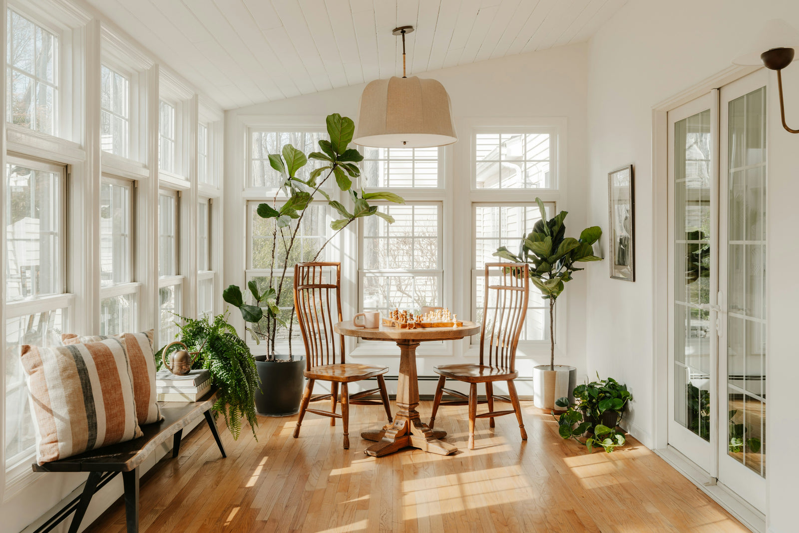 A sunlit sunroom with a chess set on a round table and plants along the windows