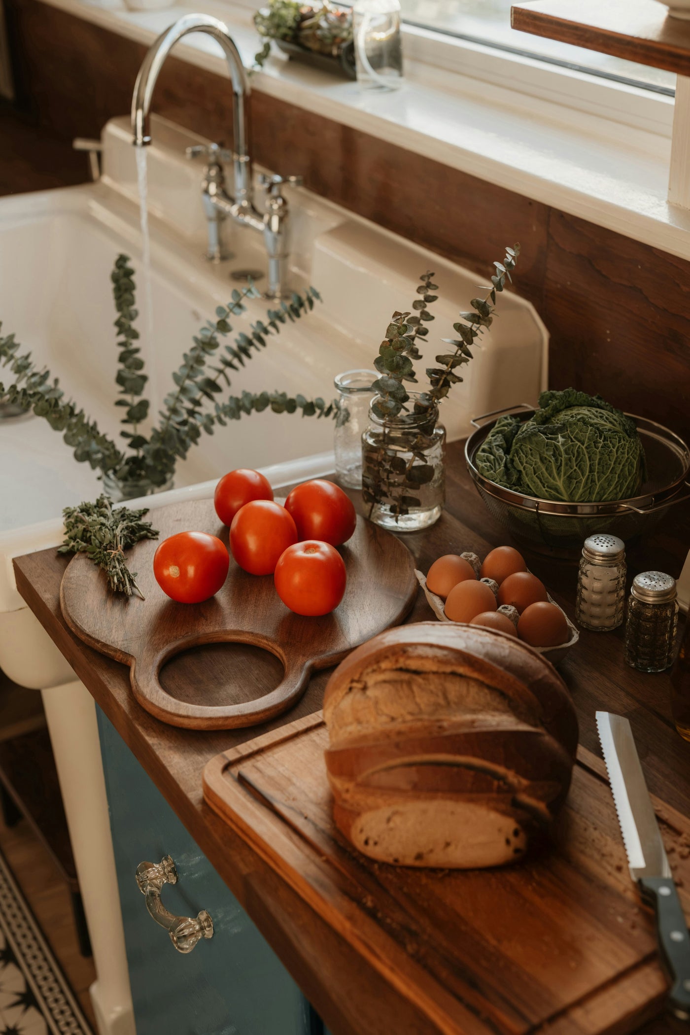 Tomatoes, herbs, eggs and a sliced loaf of bread on a wooden counter beside a farmhouse sink