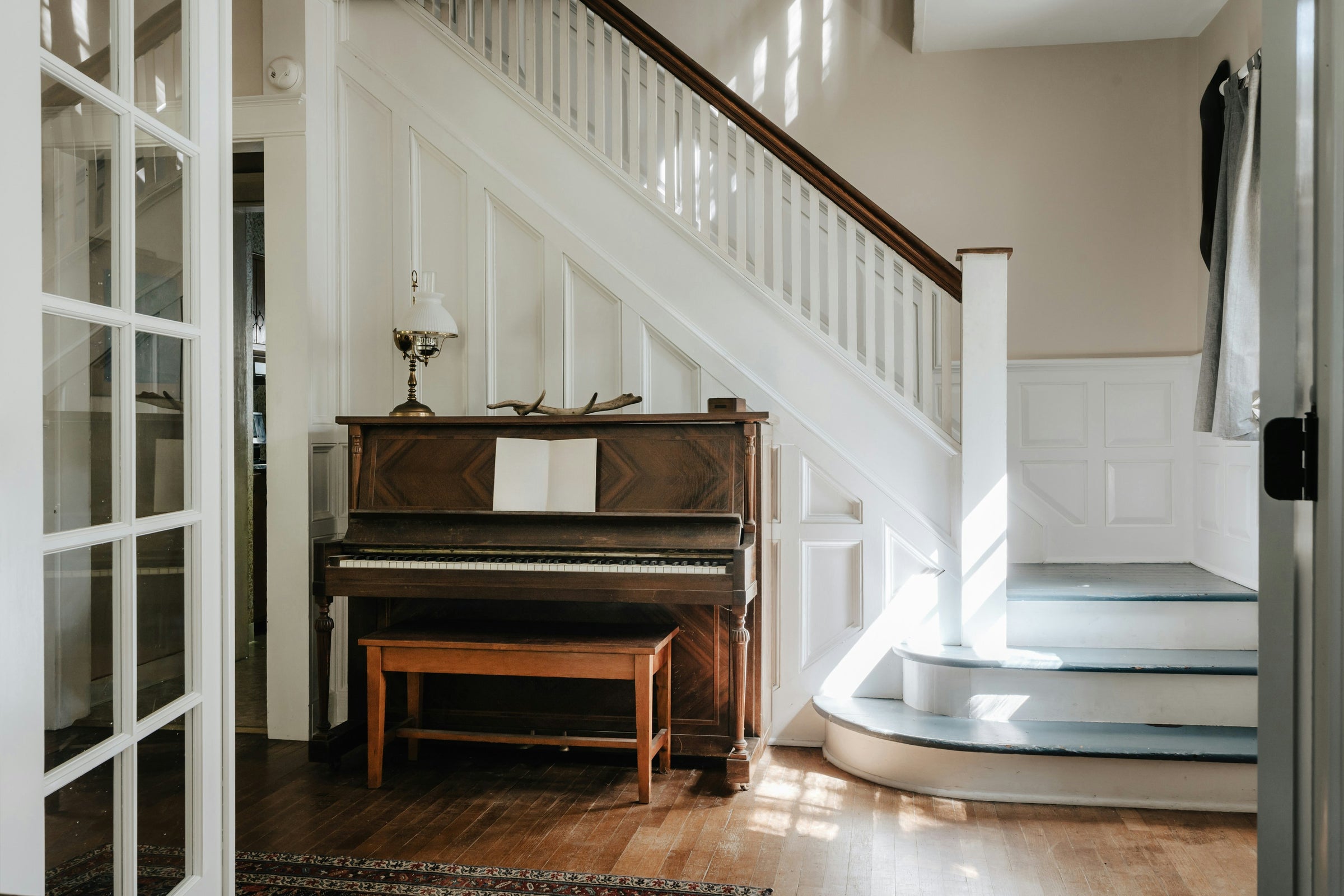 An upright piano at the foot of a staircase in afternoon light.