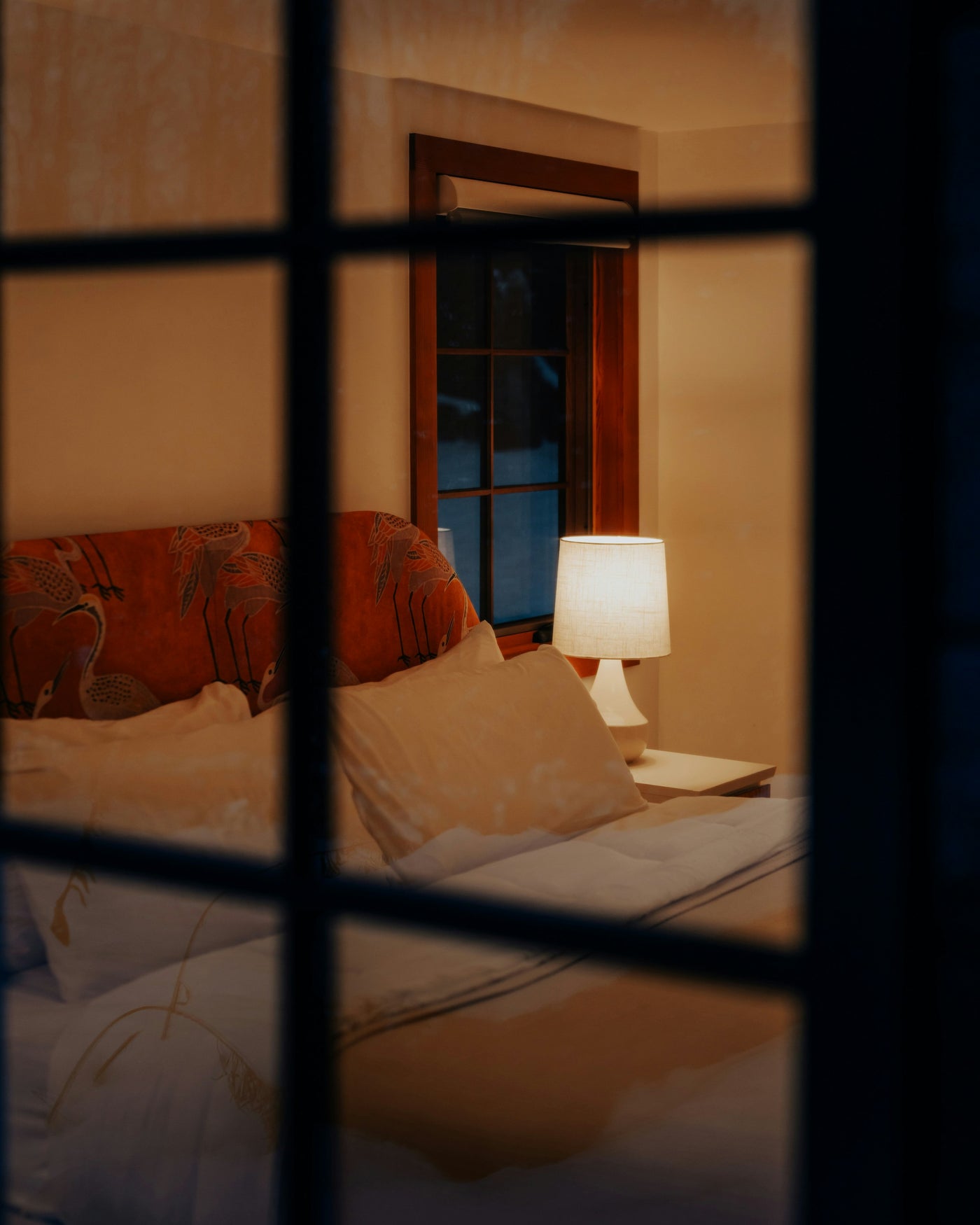 A view through a window pane into a warm bedroom, the bed made and a lamp left on, snow visible outside